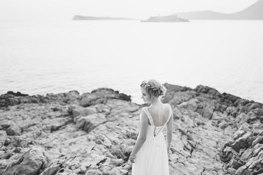 A tender bride stands on a rock by the sea, in front of her is an island, back view, black and white photo. High quality photo