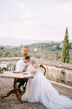The wedding couple sits at the dinner table on the roof of the old villa, the groom holds the bride's hands. Wedding at an old winery villa in Tuscany, Italy