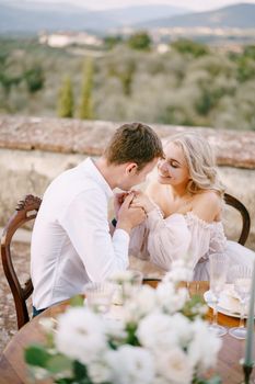 The wedding couple sits at the dinner table on the roof of the old villa, the groom holds the bride's hands. Wedding at an old winery villa in Tuscany, Italy