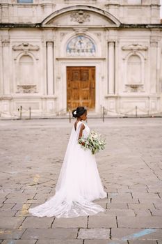 Interracial wedding couple. Wedding in Florence, Italy. An African-American bride in a white dress with a bouquet in hand, with a long veil stands in the street, against the backdrop of the church.