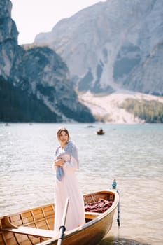 A bride stands in a wooden boat at Lago di Braies in Italy. Wedding in Europe, at Braies lake. A young girl in a white wedding dress, covers her shoulders with a blue sweater.