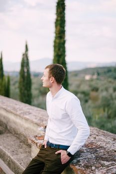 A large male portrait on the roof of an old winery villa in Tuscany, Italy. White long-sleeved shirt, smart watch, blue belt, hands in pants pockets.