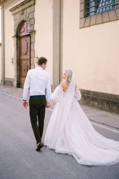 Beautiful bride and groom walking hand in hand away from the camera outside of the old villa in Italy, in Tuscany, near Florence.