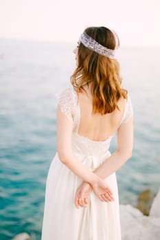 The bride stands on the seashore with her hands folded behind her back and looks into the distance. High quality photo