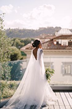 An African-American bride, with a bouquet in her hand, stands on the roof in the sunset. Destination fine-art wedding in Florence, Italy