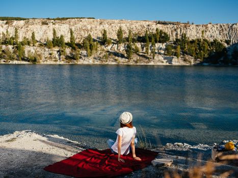 A traveler wearing a hat sits on a red cloth near the sea in nature. High quality photo