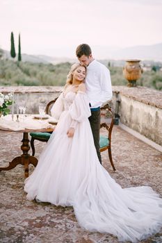 Wedding at an old winery villa in Tuscany, Italy. The wedding couple stands near the table for the wedding dinner, the groom hugs the bride at the waist.