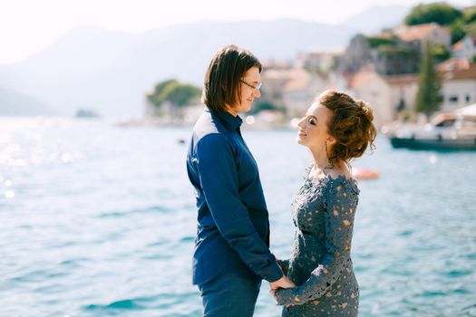 The bride in a stylish grey wedding dress and the groom stand holding hands on the seashore near the old town of Perast, close-up. High quality photo