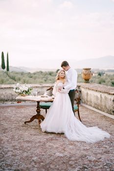 Wedding at an old winery villa in Tuscany, Italy. The wedding couple stands near the table for the wedding dinner, the groom hugs the bride at the waist.