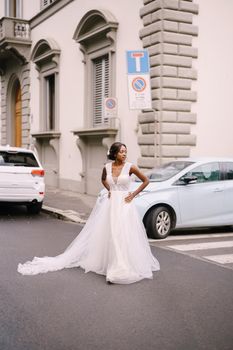 An African-American bride in a white dress, with a long veil, walks on the road on a city street. Wedding in Florence, Italy