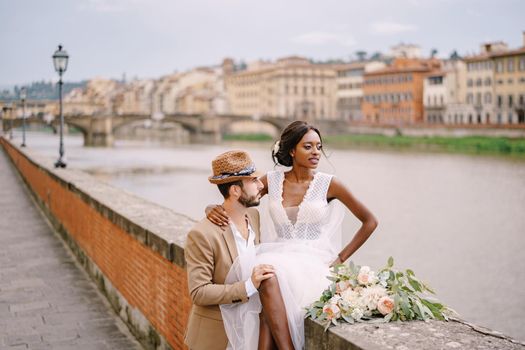 An African-American bride sits on a brick wall and a Caucasian groom hugs her. The embankment of the Arno River, overlooking city and bridges. Interracial wedding couple. Wedding in Florence, Italy.