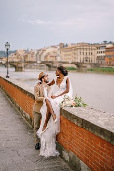 An African-American bride sits on a brick wall and a Caucasian groom hugs her. The embankment of the Arno River, overlooking city and bridges. Interracial wedding couple. Wedding in Florence, Italy.