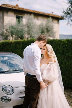 Beautiful bride and groom looking at each other and holding hands in front of a convertible at the Villa in Tuscany