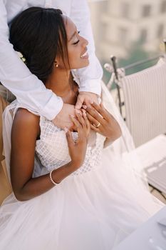 Interracial wedding couple. Destination fine-art wedding in Florence, Italy. African-American bride sits at wedding table, Caucasian groom hugs her shoulders.