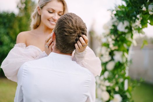 The groom holds the bride in his arms. Wedding at an old winery villa in Tuscany, Italy. Round wedding arch decorated with white flowers and greenery in front of an ancient Italian architecture.