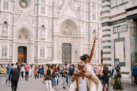 An African-American bride hopped onto the shoulders of a Caucasian groom in the Piazza del Duomo. Mixed-race wedding couple. Wedding in Florence, Italy.