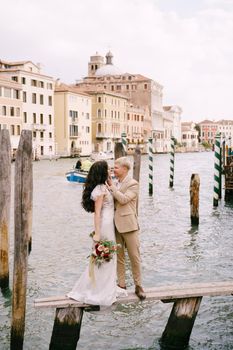 The bride and groom stand on a wooden dock for boats and gondolas, near striped green and white mooring poles, against the facades of the Grand Canal buildings.