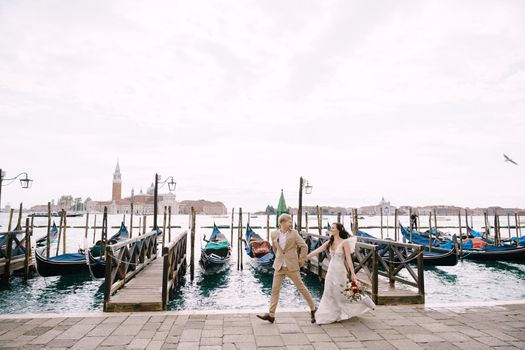 The bride and groom walk along the gondola dock holding hands in Venice, near Piazza San Marco, overlooking San Giorgio Maggiore and the sunset sky. The largest pier for gondolas in Venice, Italy.