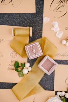Wedding rings in a box, invitation cards, silk ribbon, white roses on black background. Overhead top view