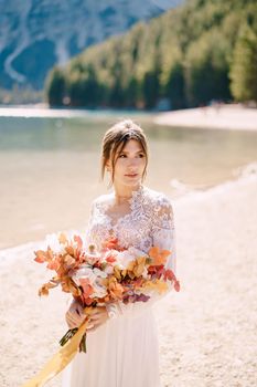 Beautiful bride in a white dress with sleeves and lace, with a yellow autumn bouquet of dried flowers and peony roses, at Lago di Braies in Italy. Destination wedding in Europe, at Braies lake.