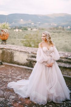 The bride in a white lush dress, with bare shoulders and puffed sleeves, walks on the roof of an old villa. Wedding at an old winery villa in Tuscany, Italy.