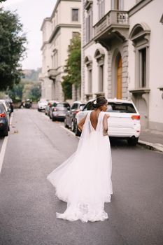An African-American bride in a white dress, with a long veil, walks on the road on a city street. Wedding in Florence, Italy