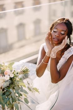 Destination fine-art wedding in Florence, Italy. African-American bride sits at the table, touches his face with her hands in gloves, a bouquet lies on the table.