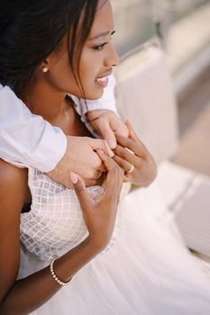 Interracial wedding couple. Destination fine-art wedding in Florence, Italy. African-American bride sits at wedding table, Caucasian groom hugs her shoulders.