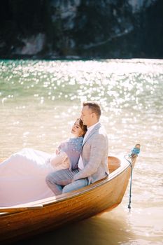 The wedding couple sits in a wooden boat at Lago di Braies in Italy. Newlyweds in Europe, at Braies Lake, in the Dolomites. The groom hugs the bride.