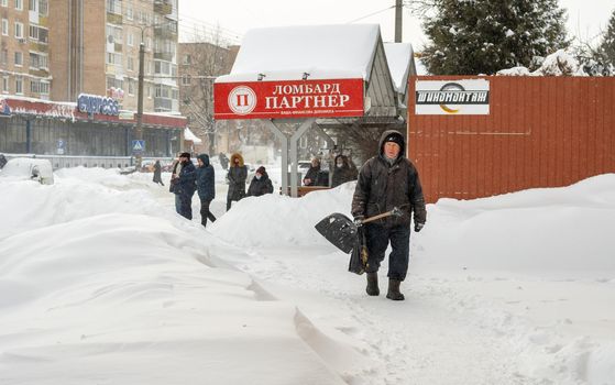 Lutsk, Ukraine - February 12,2020: City street after blizzard. An elderly man with snow shovel in winter. People walk in snow drifts on uncleaned street. Sidewalk covered in snow after storm.