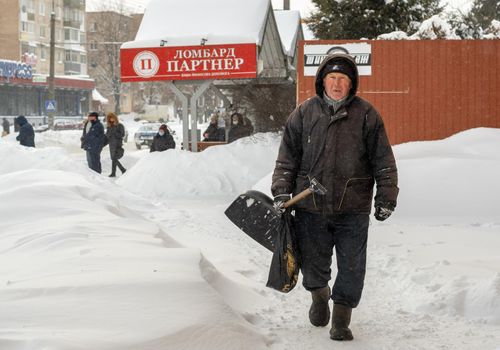 Lutsk, Ukraine - February 12,2020: City street after blizzard. An elderly man with snow shovel in winter. People walk in snow drifts on uncleaned street. Sidewalk covered in snow after storm.
