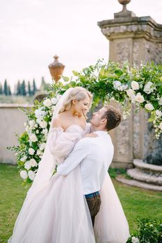 The groom is circling the bride in her arms. Round wedding arch decorated with white flowers and greenery in front of an ancient Italian architecture. Wedding at an old winery villa in Tuscany, Italy.