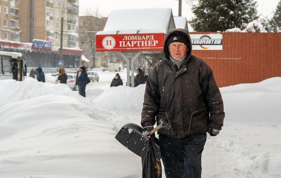 Lutsk, Ukraine - February 12,2020: City street after blizzard. An elderly man with snow shovel in winter. People walk in snow drifts on uncleaned street. Sidewalk covered in snow after storm.