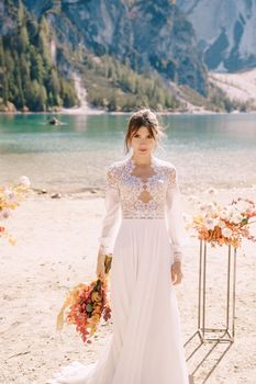 Beautiful bride in a white dress with sleeves and lace, with a yellow autumn bouquet against backdrop of arch for ceremony, at Lago di Braies in Italy. Destination wedding in Europe, at Braies lake.