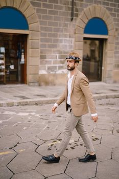 A young guy with a straw hat, a white unbuttoned shirt and a sand jacket, a small beard walks around of Florence.