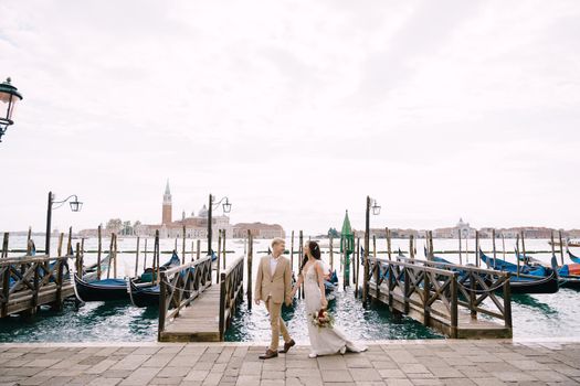 The bride and groom walk along the gondola dock holding hands in Venice, near Piazza San Marco, overlooking San Giorgio Maggiore and the sunset sky. The largest pier for gondolas in Venice, Italy.