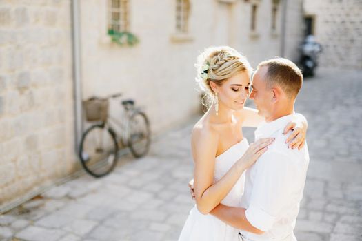 The bride and groom hug on the street of the old town of Perast next to a white building and a bicycle . High quality photo