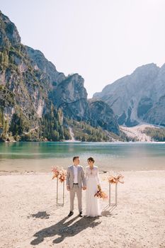 The bride and groom stand still for the ceremony, with an arch of autumn floral columns, against the backdrop of Lago di Braies in Italy. Destination wedding in Europe, at Braies lake.