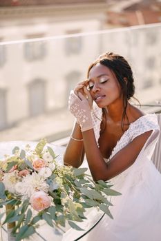Destination fine-art wedding in Florence, Italy. African-American bride sits at the table, touches his face with her hands in gloves, a bouquet lies on the table.