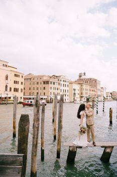 The bride and groom stand on a wooden dock for boats and gondolas, near striped green and white mooring poles, against the facades of the Grand Canal buildings.