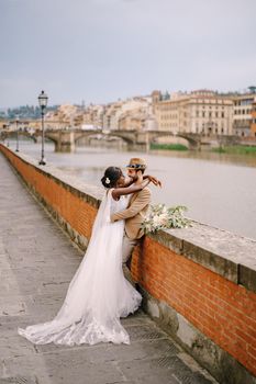 African-American bride and Caucasian groom stand hugging on the embankment of the Arno River, overlooking the city and bridges. Interracial wedding couple. Wedding in Florence, Italy.