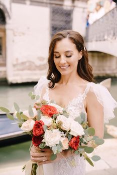 A bride in a white dress, with a train, with a bouquet of white and red roses in her hands, stands on the pier near the moored gondola in a narrow Venetian canal.