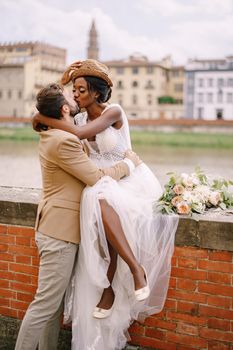 An African-American bride sits on a brick wall and a Caucasian groom hugs her. The embankment of the Arno River, overlooking city and bridges. Interracial wedding couple. Wedding in Florence, Italy.