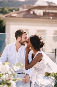 Interracial wedding couple. Destination fine-art wedding in Florence, Italy. African-American bride and Caucasian groom are sitting at rooftop wedding dinner table overlooking the city.