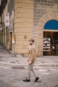 A young guy with a straw hat, a white unbuttoned shirt and a sand jacket, a small beard walks around of Florence.