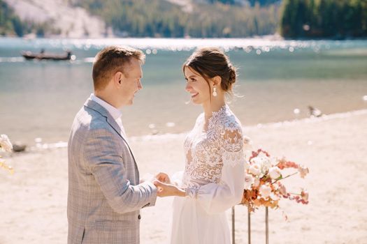 The bride dons a ring to the groom at the venue for the ceremony, with an arch of autumn floral columns, against the backdrop of Lago di Braies in Italy. Destination wedding in Europe, at Braies lake.