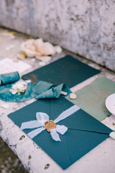 Blue mockup of invitations to wedding, blue envelope with seal print of sealing wax, a silk blue ribbon, a white sheet with the colors of dess code, pink flower against background of marble stone