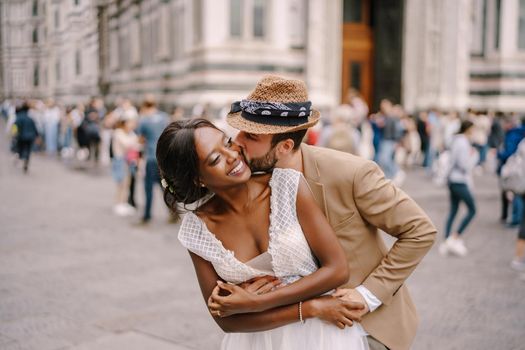 Caucasian groom hugs from behind and kisses African-American bride. Multiethnic wedding couple. Wedding in Florence, Italy.
