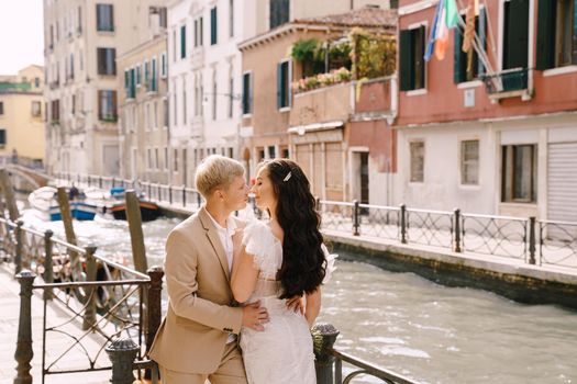 Newlyweds stand hugging on the banks of the Venice Canal. The groom hugs the bride by the waist. White wedding dress with a small beautiful train and a sandy color men's suit.
