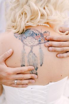 Bride with dreamcatcher tattoo on her back, groom hugs the bride and puts his hands on her back, close-up . High quality photo